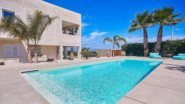 A modern pool area with a clear blue pool, a two-story building with balconies, and palm trees. - VILLA CIURIDDA (Alcamo)