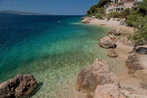 A coastal area with clear turquoise waters, rocky shoreline, and buildings on the right.