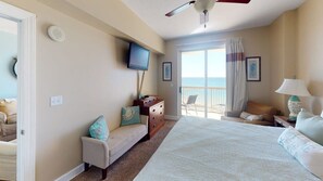 Un dormitorio con cama, cómoda, televisor, ventilador de techo, ventana con vistas y una silla junto a la ventana.