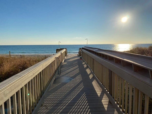 Ein Holzsteg führt zum Strand, der Himmel ist klar und die Sonne scheint.