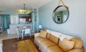 A living room with a beige sofa, a dining table with chairs, a kitchen area, and a large mirror on the wall. - Coastal Condo 807 | Beachfront! (Panama City)