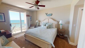 A bedroom with a large bed, a ceiling fan, a view of the ocean through a sliding door, and a small table with a lamp.