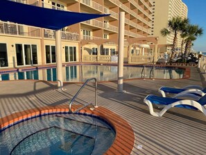 Zona de piscina amb banyera d'hidromassatge, gandules i un edifici al fons amb balcons.
