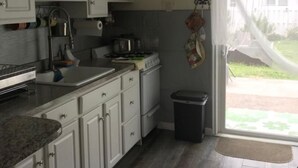 A kitchen with white cabinets, a stove, a sink, and a trash bin. There is a window with a view of the outside.