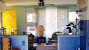 A compact kitchen with a central island, a dining area with a red chair, and a living room with a sofa and a lamp.