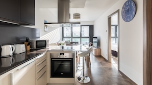 Una cocina moderna con horno empotrado, microondas y hervidor, un comedor con taburete de bar, y un ventanal que proporciona luz natural.