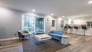 A modern living room with a large window, a sofa, a coffee table, and a kitchen area with bar stools. - 6807 Jones Valley · ֍The Strata֎ 4000 SQ Feet, 6 BR, Pool Table (Huntsville)