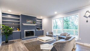 A modern living room with a fireplace, grey sofas, and a large window with a view of trees. - 6807 Jones Valley · ֍The Strata֎ 4000 SQ Feet, 6 BR, Pool Table (Huntsville)