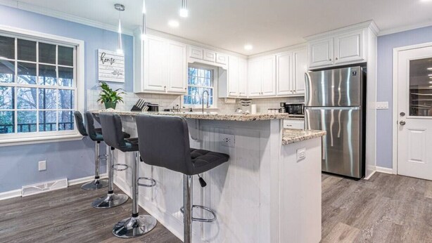 A modern kitchen with white cabinets, a stainless steel refrigerator, and a bar area with two stools. - 6807 Jones Valley · ֍The Strata֎ 4000 SQ Feet, 6 BR, Pool Table (Huntsville)