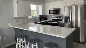 A modern kitchen with white cabinets, stainless steel appliances, a large island with stools, and a window with a view. - 2 blocks to the beach- Pool- King-sized beds- Sound- SUPs- Linens- Beachgear (North Topsail Beach)