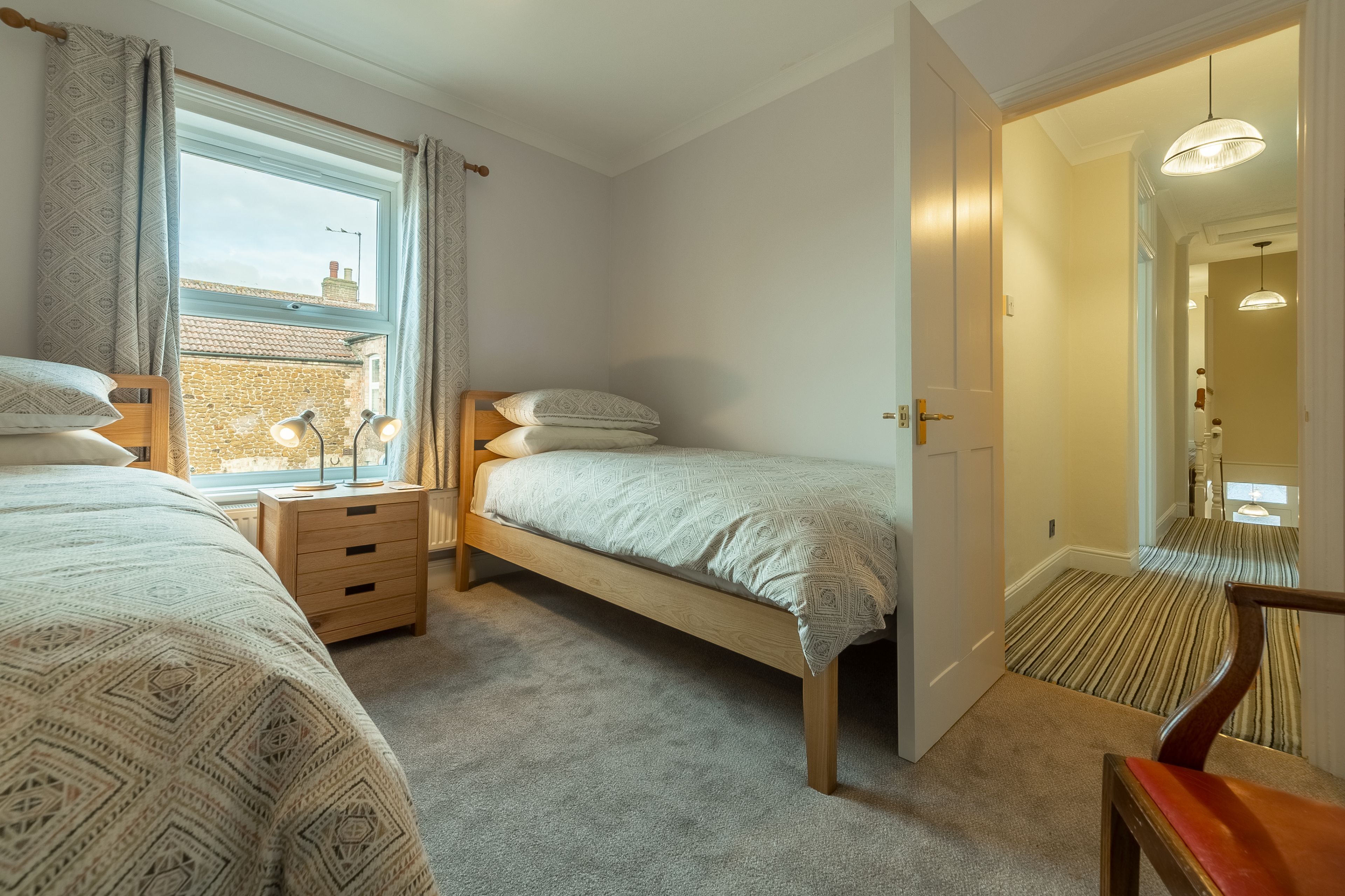 A bedroom with two beds, a wooden nightstand, a bedside table with lamps, and a view of the outside through a window.
