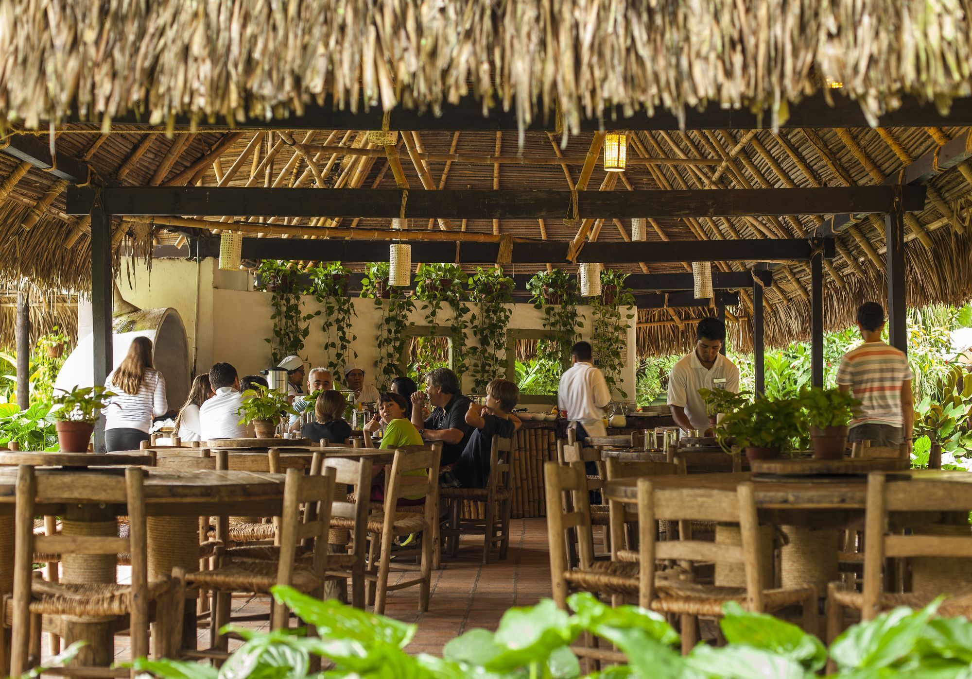 Un restaurante al aire libre con mesas y sillas de madera, techos de paja y plantas en macetas.