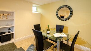 A dining area with a glass table, black chairs, a wine bottle, and glasses on the table. There is a shelf with decorative items and a mirror on the wall. - Anns Cottage, Chapel Stile (Ambleside)