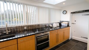 A modern kitchen with a black countertop, wooden cabinets, a built-in oven, and a window with blinds. - Anns Cottage, Chapel Stile (Ambleside)