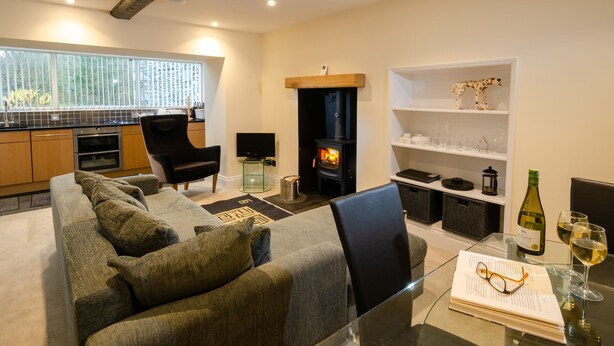 A modern living room with a sofa, a fireplace, a kitchen area, and a dining table with glasses and a bottle. - Anns Cottage, Chapel Stile (Ambleside)