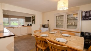 Zona pranzo con tavolo e sedie in legno, cucina con mobili bianchi e vista sull'esterno attraverso le finestre.