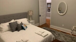 A bedroom with a bed, pillows, a nightstand, a lamp, a mirror, and a door leading to another room.