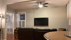A living room with a brown leather sofa, a flat-screen TV mounted on the wall, a ceiling fan, and a wooden dining table.