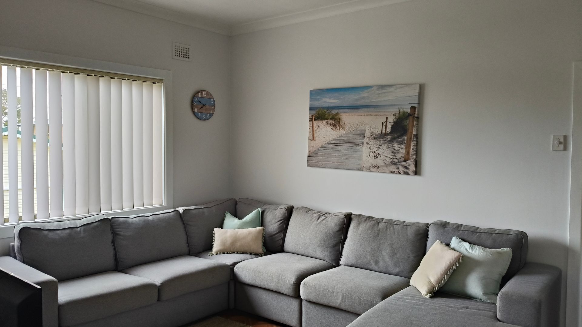 Un salon avec un canapé d’angle gris, une horloge murale et un tableau représentant une scène de plage.