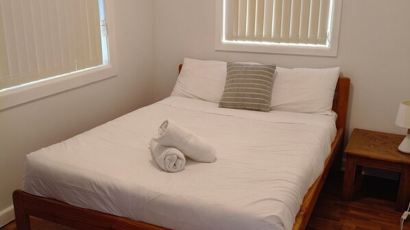 A neatly made bed with white linens, a rolled towel, and two pillows. There is a small wooden nightstand with a lamp beside the bed.
