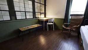Una habitación con piso de madera, un escritorio con silla, una silla de mimbre y un banco.