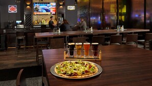 Un espace bar moderne avec un téléviseur diffusant des événements sportifs, un comptoir avec des bouteilles et une pizza sur une table.