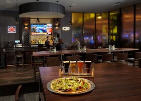 Zona de bar moderna con una televisión mostrando deportes, una barra con botellas y una pizza sobre una mesa.