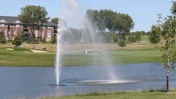 A golf course with a fountain in the middle of a lake, surrounded by a building with multiple windows and a lawn.