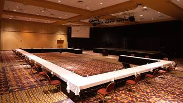 A large conference room with a long table, rows of chairs, and a stage with a screen.