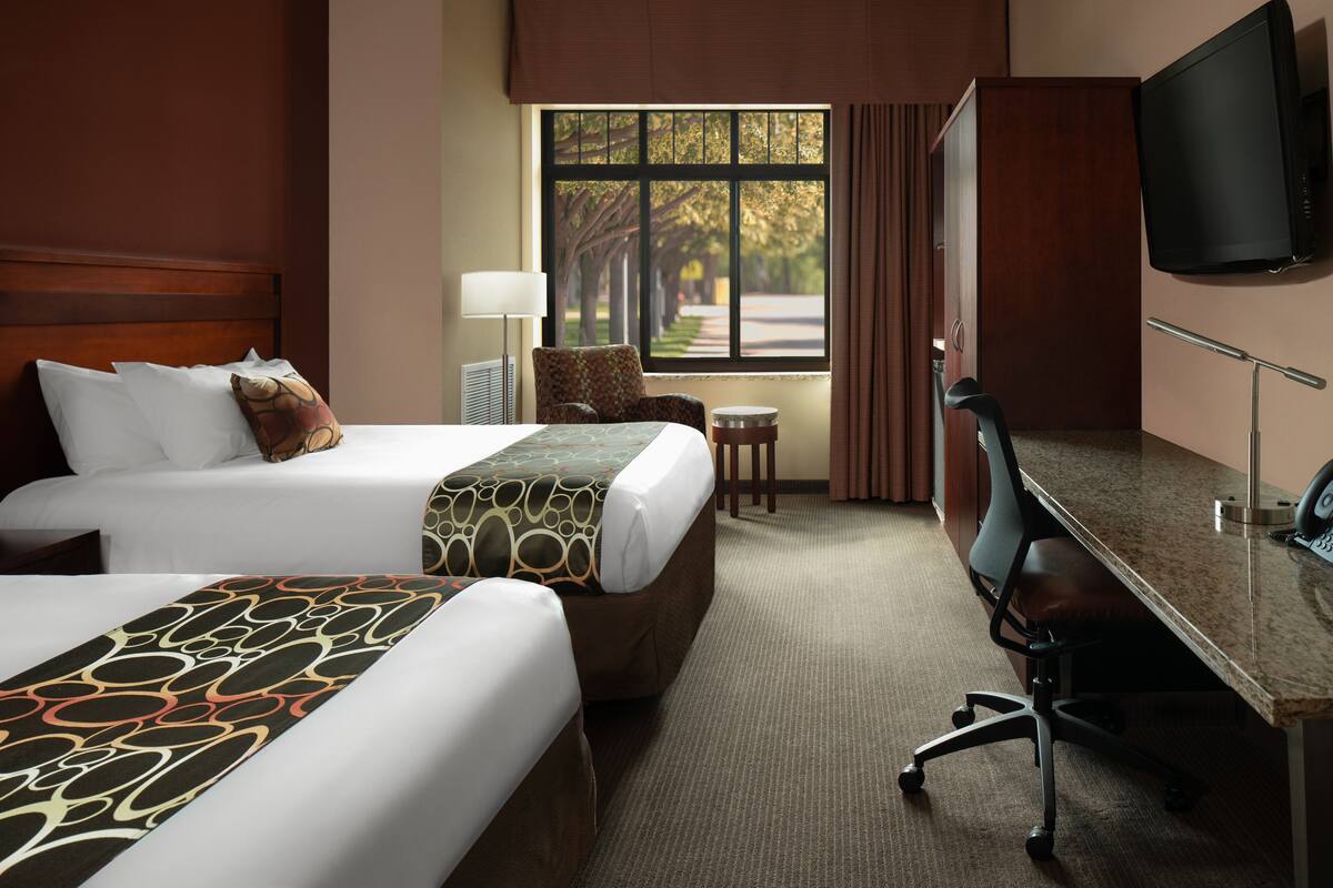 A hotel room with two beds, a desk, a chair, a TV, and a window with a view of trees.