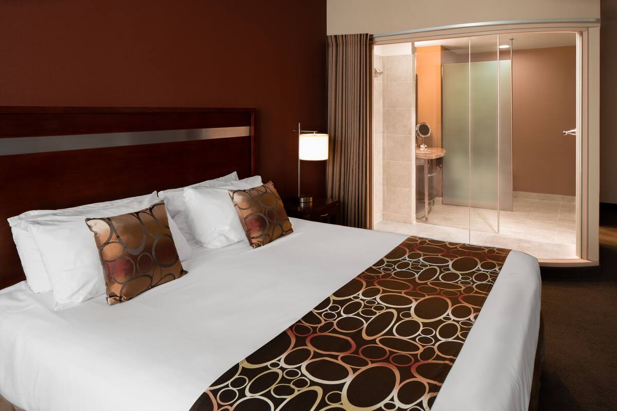 A hotel room with a large bed, a bedside table with a lamp, a bathroom with a glass shower enclosure, and a mirror.