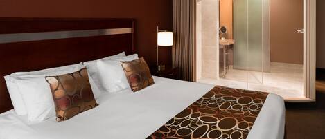 A hotel room with a large bed, a bedside table with a lamp, a bathroom with a glass shower enclosure, and a mirror.