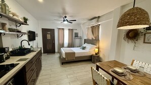 Ein kompaktes Hotelzimmer mit Küchenzeile, einem Himmelbett, einer Essecke mit Tisch und Stühlen sowie einem Deckenventilator.