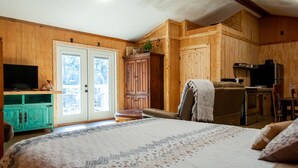A bedroom with a large bed, wooden walls, a television, a cabinet, and a kitchen area. - studio rm w/ full kitchen, 1 bth , private deck, & WiFi @ Kick Back Ranch. (Ramer)