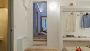 A hotel room with a stone floor, a bed with a white cover, a table with two pomegranates, a hanging clothing rack with two shirts, and a staircase leading to another room. - Masseria Pugliese Deluxe (Ostuni)