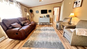 Un salon avec un canapé en cuir marron, un fauteuil gris, une table basse en bois, un tapis gris, une armoire blanche et un téléviseur à écran plat fixé au mur.