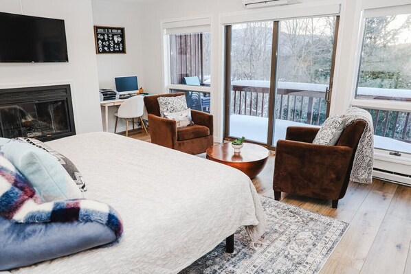 A cozy bedroom with a large bed, two brown armchairs, a small table, and a desk with a laptop. A fireplace is visible, and there are large windows offering a view of a snowy landscape.