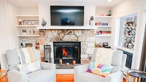A living room with a stone fireplace, two grey armchairs, a television above the fireplace, and a bookshelf with various items.