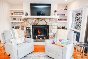 Un salón con chimenea de piedra, dos sillones grises, un televisor sobre la chimenea y una estantería con diversos objetos.