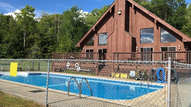 A wooden cabin with a swimming pool surrounded by a chain-link fence.