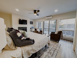 A bedroom with a large bed, a ceiling fan, a fireplace, a desk with a chair, and large windows with a view of the outdoors.
