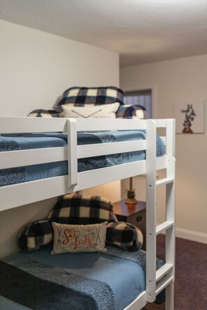 Een stapelbed met een geruit dekbed en kussens, een kleine tafel met een lamp en een lijst met een foto aan de muur.
