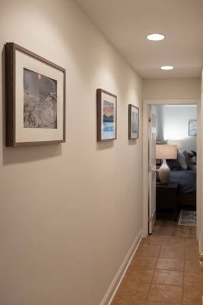 A hallway with three framed pictures on the wall, leading to a bedroom with a bed and a nightstand.
