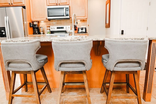 Una cocina con una isla central, tres taburetes de bar, un microondas, una cafetera y un refrigerador.
