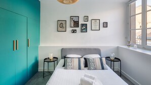 A modern bedroom with teal walls, a grey headboard, and a bed with patterned pillows. There are framed pictures on the wall, a window with a view, and a small table with a lamp.