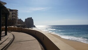 A curved walkway along a beach with a rocky cliff and buildings in the background. - Private Oceanfront Luxury (Cabo San Lucas)