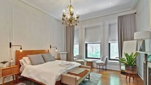Un dormitorio con una cama grande, mesitas de noche, un banco, una araña, una silla, una planta y una chimenea.