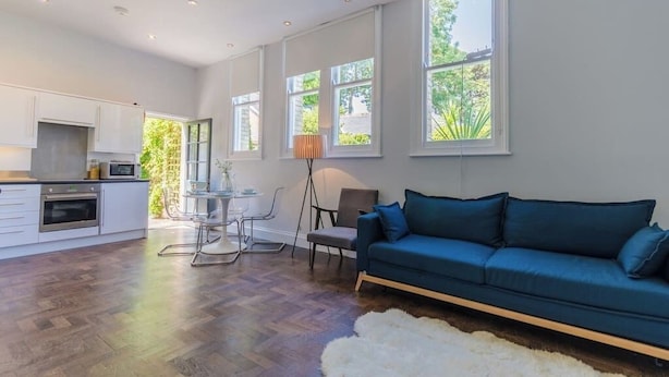 A spacious living area with a sofa, a dining table, and chairs. There is a kitchen area with cabinets and appliances. Large windows provide natural light and views of greenery outside. - Charming Schoolhouse Apartment by Fowey Harbour (Cornwall)