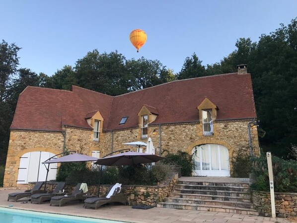 A stone building with a swimming pool, lounge chairs, and a hot air balloon in the sky.