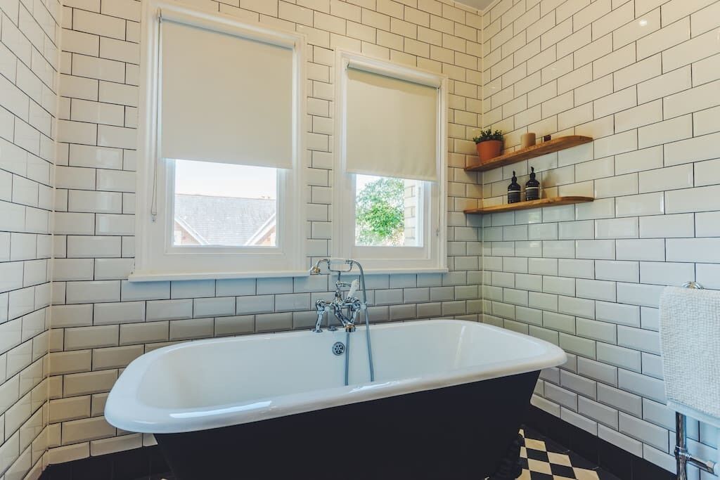 Un baño con una bañera independiente, paredes de azulejos blancos y dos ventanas con persianas blancas.